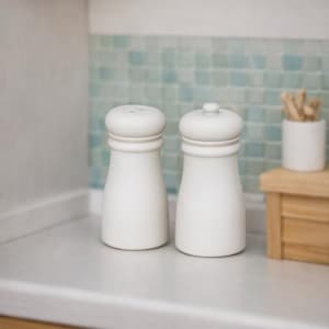 May include: Two off-white wooden salt and pepper shakers with rounded tops and a classic design. One shaker has small holes on top. The shakers sit on a white surface, with a light blue tiled backsplash in the background.