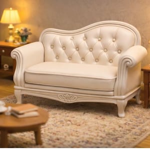 Op de afbeelding: Een crèmekleurige miniatuur loveseat met een gecapitonneerde rug en opgerolde armen. De kleine bank heeft een decoratieve gesneden schort en staat op een patroonkleed. Een kleine ronde tafel en lampen staan op de achtergrond.