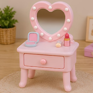 May include: A light pink vanity table featuring a heart-shaped mirror with integrated white lights. The tabletop holds a small open compact, a lipstick tube, and a small cylindrical object. The vanity has a single drawer with a round knob.