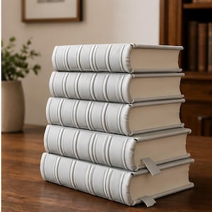 May include: A stack of five hardcover books with light gray covers and cream-colored pages. The spines have a ribbed design. A silver bookmark is inserted in one of the books. The books are stacked on a wooden surface.