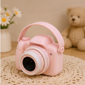 May include: A light pink toy camera featuring a rounded body, a prominent central lens with a white rim, a top-mounted carrying handle, and a shutter button on the right side. The device has a smooth, matte finish and a compact, portable design.