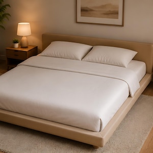 Op de afbeelding: Een beige bed met bijpassend hoofdbord en frame is opgemaakt met een lichtbeige lakenset en twee kussens. Een klein houten nachtkastje met een lamp en een kleine plant staat links van het bed.