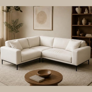 Könnte beinhalten: Ein großes, cremefarbenes Ecksofa mit modernem Design. Das Sofa hat eine niedrige Form, eckige Armlehnen und schwarze Metallbeine. Es steht in einem neutral gehaltenen Wohnzimmer mit einem runden Couchtisch aus Holz und einem gerahmten Kunstwerk.