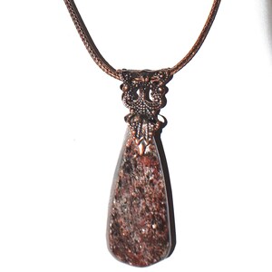 Puede incluir: Un collar con colgante en forma de lágrima que presenta una piedra moteada en tonos marrones y negros, engarzada en un adorno de metal con acabado en cobre envejecido. El colgante cuelga de un cordón trenzado fino de color marrón.