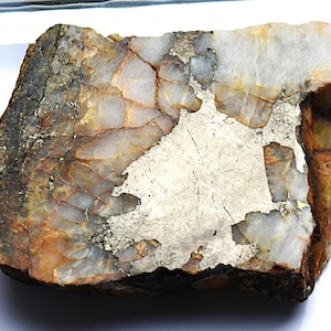 May include: A rough-cut stone with a mix of colors, including white, brown, and gold. The stone has a crystalline structure with visible fractures and a metallic sheen in some areas. The overall shape is irregular and natural.