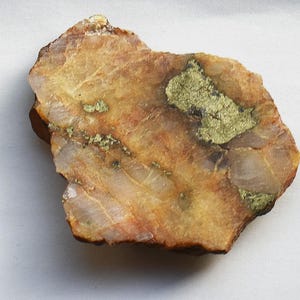 May include: A rough-hewn rock sample with a mix of colors, including tan, brown, and gold. The rock has a layered appearance with visible veins of gold-colored mineral deposits. The overall texture is coarse and natural, suggesting a raw, unrefined state.