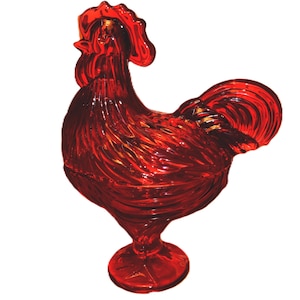 May include: A translucent red glass rooster figurine standing on a circular pedestal base. The glass features molded details representing feathers on the body and tail, with a distinct comb on the head, all rendered in a vibrant, deep red color.