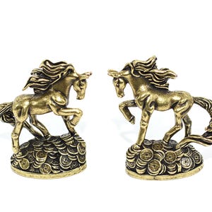 May include: Two brass-colored metal horse figurines, each depicted in a dynamic rearing pose. The horses are standing on a base composed of a pile of circular coins with square center holes, featuring a weathered, antique metallic finish.