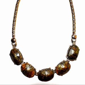 May include: A necklace featuring five oval-shaped, dark brown and green stones. The stones are connected by silver-toned metal wire and are strung on a silver-toned chain. The stones have a speckled appearance.
