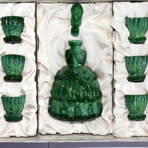 May include: A vintage green glass decanter set, including a bottle shaped like a woman's dress and six matching shot glasses. The set is nestled in a white satin-lined box, offering a classic and elegant presentation. The glass has a textured design.