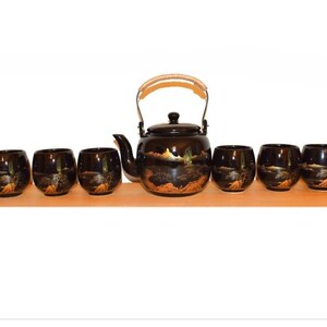 May include: A black tea set consisting of one teapot with a curved spout, a lid, and a woven handle, accompanied by six matching tea cups. Each piece features a hand-painted gold landscape design depicting mountains, trees, and rocky terrain.