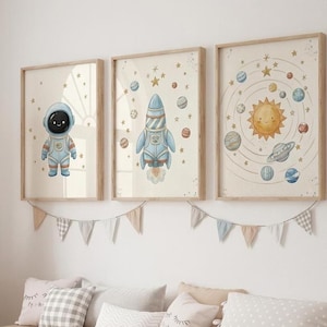 May include: Three framed art prints with a space theme. The prints feature an astronaut, a rocket, and a solar system illustration. The artwork is in soft pastel colors, with a wooden frame. A decorative banner hangs below the prints.