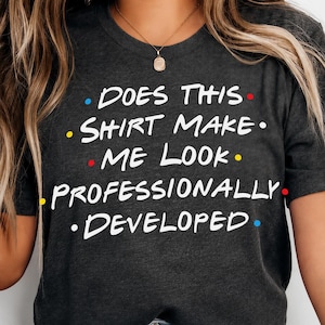 May include: A dark charcoal grey short-sleeve t-shirt featuring the white text "DOES THIS SHIRT MAKE ME LOOK PROFESSIONALLY DEVELOPED" with small blue, red, and yellow dots interspersed between the words in a multi-line graphic design.