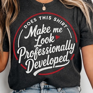 May include: A dark heather grey short-sleeve t-shirt featuring a red circular graphic with white text that reads: DOES THIS SHIRT Make me Look Professionally Developed, including a small red heart icon between the words Look and Professionally.