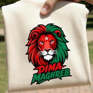 May include: A cream-colored t-shirt featuring a graphic of a lion's head with a split red and green mane. A white five-pointed star is centered on the lion's forehead. Below the lion, the text DIMA MAGHREB is printed in red and green letters.