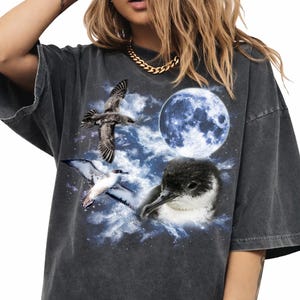 May include: Dark grey oversized t-shirt with a graphic of birds, a full moon, and clouds. The design includes a flying seagull, a bird with white and black wings, and a small black and white bird. The shirt has a vintage, washed appearance.