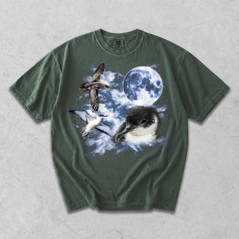 May include: Green t-shirt with a graphic design of birds, a full moon, and clouds. The design includes a detailed illustration of a bird in flight, a close-up of a bird's head, and a full moon in a blue sky.