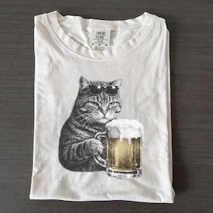 以下が含まれることがあります： サングラスをかけた猫がビールジョッキを持っている白クリーム色のTシャツ。ビールには泡があります。シャツの首元にはComfort Colorsのタグが付いています。