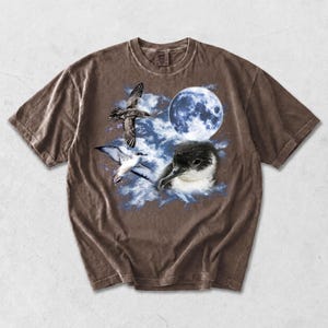 May include: Brown t-shirt with a graphic of a full moon, birds, and a bird's head. The design includes a blue moon, white clouds, and detailed illustrations of birds in flight. The shirt has a vintage, worn appearance.