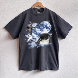 May include: Dark grey t-shirt with a graphic of seabirds, a full moon, and clouds. The design includes a close-up of a bird's head and two birds in flight. The shirt is hanging on a wooden hanger.