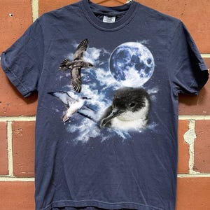 May include: Navy blue t-shirt with a graphic of birds, a full moon, and clouds. The design includes a flying bird, a bird with its wings spread, and a bird's head. The shirt is hanging on a metal hanger against a brick wall.