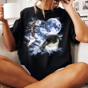 May include: Black t-shirt with a celestial design featuring a full moon and three birds. The graphic includes a flying bird, a bird with white and grey feathers, and a dark bird with a white beak. The design is set against a cloudy, blue background.