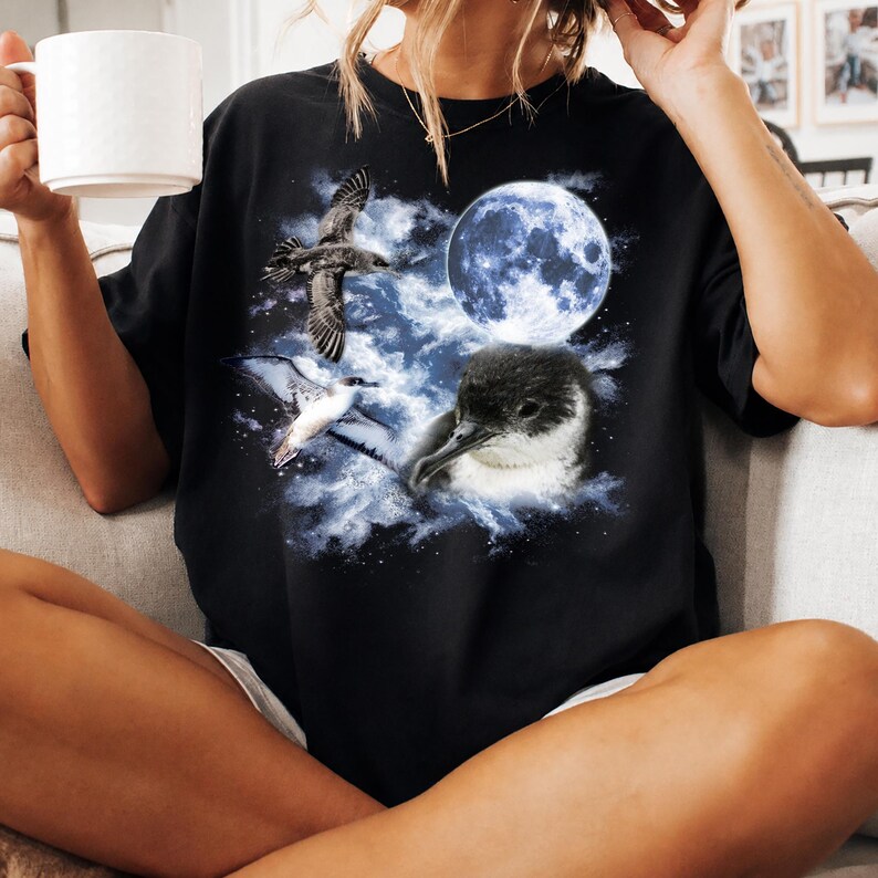 May include: Black t-shirt with a celestial design featuring a full moon and three birds. The graphic includes a flying bird, a bird with white and grey feathers, and a dark bird with a white beak. The design is set against a cloudy, blue background.