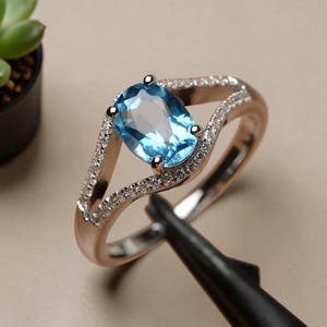 May include: A ring featuring a central oval-cut light blue gemstone held by four prongs. The band is crafted from a metallic material and features a split-shank design adorned with small, clear, sparkling stones along the upper edges.