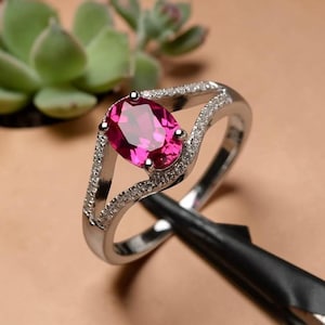 Peut inclure: Une bague argentée ornée d'une pierre précieuse rose vif de taille ovale au centre. L'anneau présente un design à double branche, chacune incrustée de petites pierres claires et scintillantes le long de la monture.