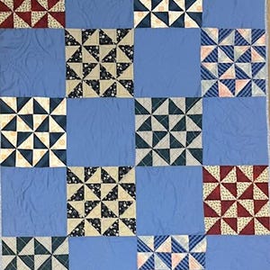 May include: A handmade quilt with a light blue background and a patchwork design. The patchwork squares feature various patterns, including triangles in navy, cream, and red with white accents. The quilt is a cozy, decorative textile.