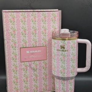 May include: A Stanley LoveShackFancy tumbler with a handle and a clear lid, featuring a pink and green floral rose pattern on a white background. The tumbler is displayed next to a matching rectangular box with the text Stanley LoveShackFancy.