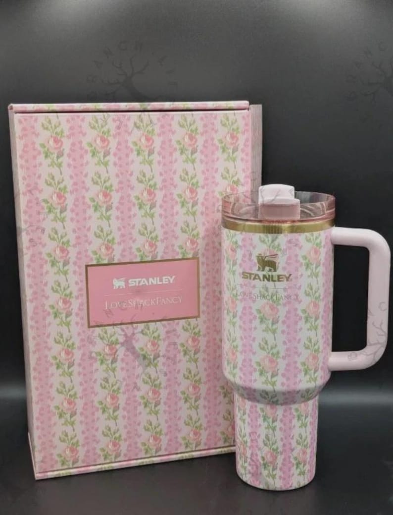 May include: A Stanley LoveShackFancy tumbler with a handle and a clear lid, featuring a pink and green floral rose pattern on a white background. The tumbler is displayed next to a matching rectangular box with the text Stanley LoveShackFancy.