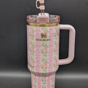 May include: A pink Stanley tumbler with a floral rose pattern and vertical stripes. The cup features a gold logo, a LoveShackFancy inscription, a light pink handle, a clear lid, and a straw topper shaped like a pink bow on a clear straw.