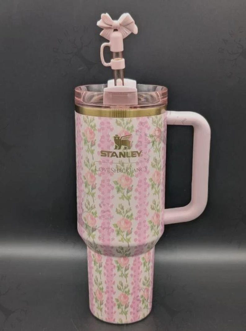 May include: A pink Stanley tumbler with a floral rose pattern and vertical stripes. The cup features a gold logo, a LoveShackFancy inscription, a light pink handle, a clear lid, and a straw topper shaped like a pink bow on a clear straw.