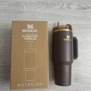 May include: A brown Stanley Flowstate tumbler with a handle and straw, next to its cardboard box. The box features the Stanley logo and product details, including "40 oz". The tumbler is designed to keep drinks cold for 2 days and hot for 7 hours.