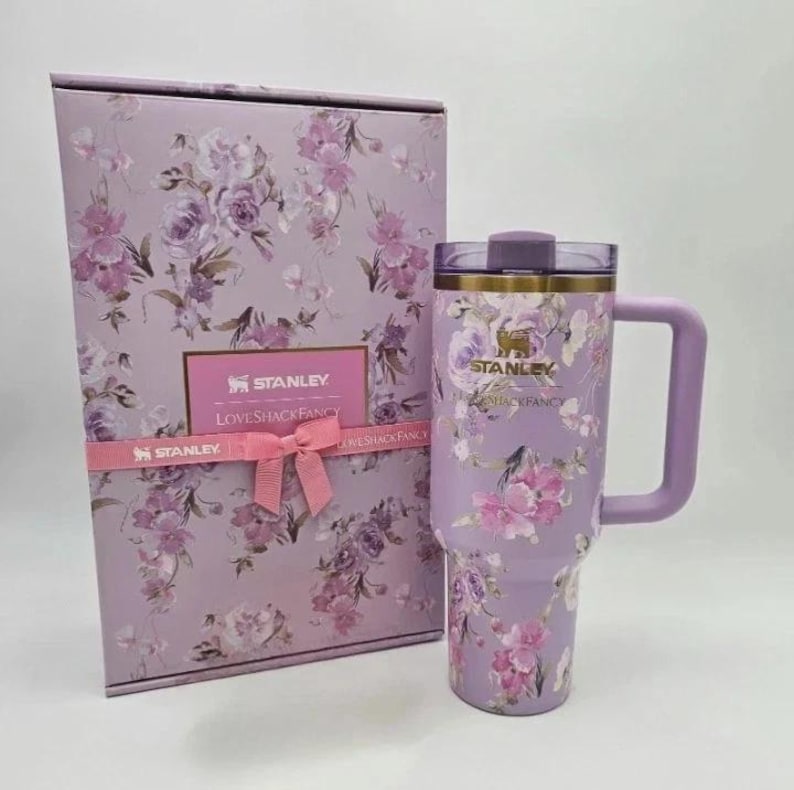 May include: A lavender Stanley tumbler with a floral design, a matching handle, and a lid. The tumbler is paired with a gift box featuring a similar floral pattern and a pink ribbon. The box and tumbler have the text "Stanley" and "LoveShackFancy".