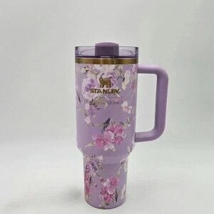 May include: A lavender Stanley tumbler with a floral design featuring pink and purple flowers. The tumbler has a handle, a lid, and the Stanley and Shackfancy logos. The tumbler is designed to keep drinks cold or hot.