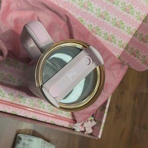 May include: A light pink insulated tumbler with a matching lid and a gold-toned rim. The tumbler features a sturdy handle and is presented inside a box lined with pink floral patterned paper and soft pink fabric, viewed from a top-down angle.