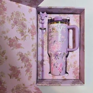 May include: A floral-patterned Stanley tumbler and straw set in a gift box. The tumbler is lavender with pink and purple flowers. The box and straw have matching floral designs. The tumbler has a handle and a gold-colored rim.