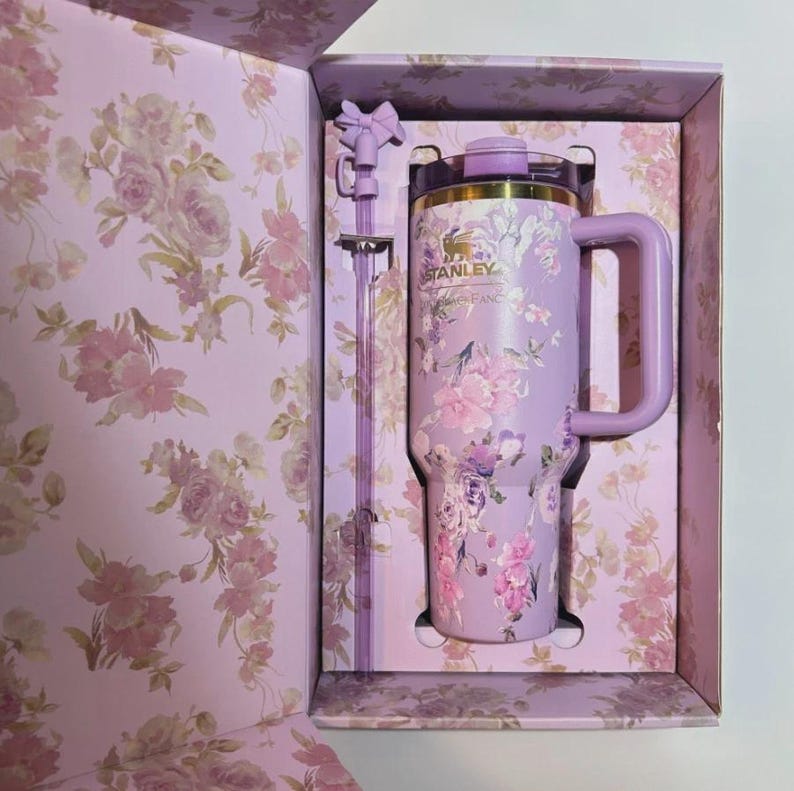 May include: A floral-patterned Stanley tumbler and straw set in a gift box. The tumbler is lavender with pink and purple flowers. The box and straw have matching floral designs. The tumbler has a handle and a gold-colored rim.
