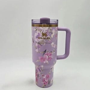 May include: A lavender Stanley tumbler with a floral design featuring pink and purple flowers. The tumbler has a handle, a lid, and the Stanley and Shackfancy logos. The tumbler is approximately 10 inches tall.