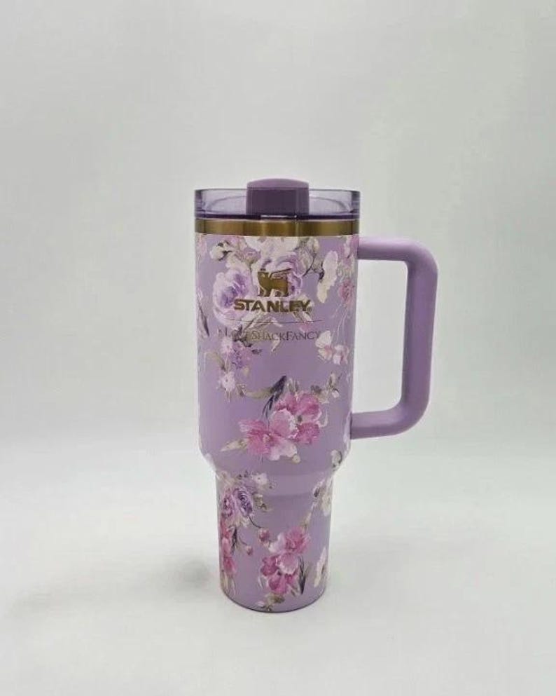 May include: A lavender Stanley tumbler with a floral design featuring pink and purple flowers. The tumbler has a handle, a lid, and the Stanley and Shackfancy logos. The tumbler is approximately 10 inches tall.