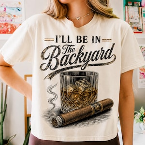 May include: A cream-coloured t-shirt featuring the phrase "I'll Be In The Backyard" in a vintage style. The graphic depicts a glass of whiskey with ice and a lit cigar. The design is a detailed illustration with a relaxed, classic theme.