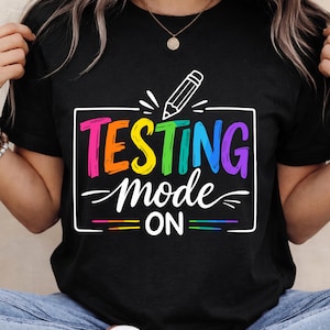 May include: Black t-shirt featuring the text "TESTING mode ON" in a rainbow gradient, with a pencil illustration. The casual shirt is a simple design, suitable for everyday wear.