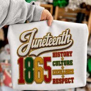 Può includere: Felpa bianca con la scritta "Juneteenth" in corsivo con dettagli oro e bianchi. Il numero "1865" è visualizzato in rosso, verde e oro. Sono visibili anche le parole "Storia, Cultura, Eccellenza, Rispetto".