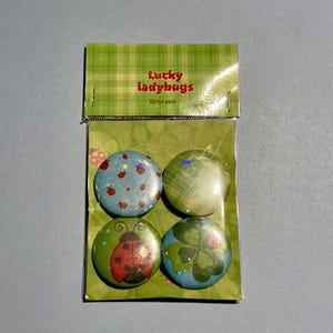 May include: A pack of four circular pin-back badges in a clear plastic sleeve with a green plaid header labelled Lucky ladybugs Badge pack. The badges feature designs of small red ladybirds on blue, a green wash, a large ladybird, and a green clover.