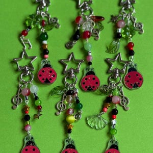 May include: Six keychains featuring red ladybird charms with black spots, attached to silver star-shaped clasps. Each keychain includes a variety of colourful beads in shades of green, red, pink, and black, along with silver music note and leaf charms.
