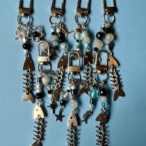 May include: Six silver-toned metal keychains featuring fish skeleton charms, star charms, and various beads in shades of blue, black, white, and clear. Each keychain is attached to a silver lobster clasp, creating a collection of dangling accessories.