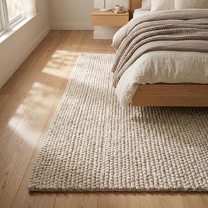 May include: A cream-coloured, chunky-knit rug in a bedroom setting. The rug is rectangular and has a textured, looped surface. The rug is placed on a light-coloured wooden floor, near a bed with a wooden frame and neutral-coloured bedding.