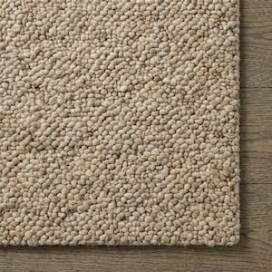 May include: Close-up of a beige textured rug with a nubby, looped pile. The rug's edge is visible, resting on a wooden floor. The rug's texture is dense and uniform, creating a soft, neutral surface.